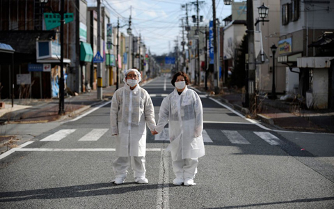 Wearing white protective masks and suits, Yuzo Mihara and his wife Yuko pose for photographs on a deserted street in the town of Namie, inside the Fukushima nuclear disaster exclusion zone. Following the 11 March 2011 earthquake, tsunami and nuclear disaster, tens of thousands of people lost their homes and are still living in temporary housing. The 21,000 residents of Namie had to abandon their homes after the town was evacuated following the nuclear alert.