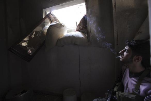 Chiến binh phiến quân ngồi hút thuốc trong khi theo dõi 	quân chính phủ qua một chiếc gương vỡ trong căn phòng ở Deir al-Zor, Syria.