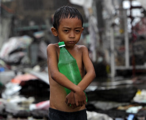 Một cậu bé ôm chai nước trong khi cởi trần đi dưới trời mưa giữa những đống đổ nát tại thành phố Tacloban, Philippines.