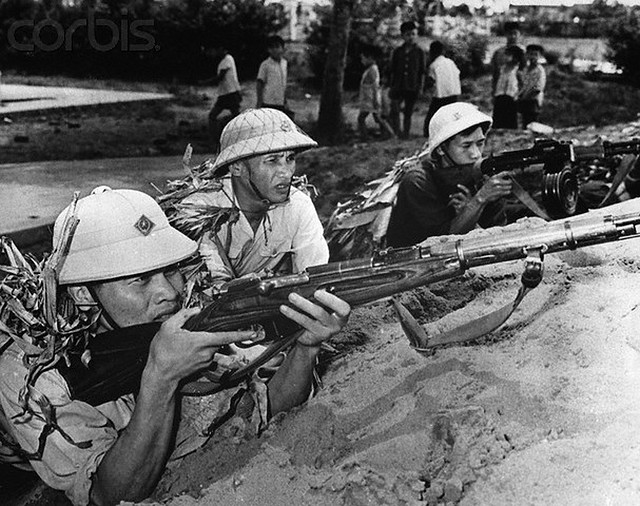 7/16/1965-Hanoi, North Vietnam: Children play nearby as members of a North Vietnamese self-defense team train. Many such teams and militia units have been formed in North Vietnam. --- Image by © Bettmann/CORBIS