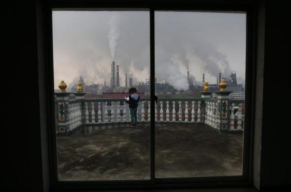 Một bé gái đọc sách trên ban công khi khói bốc lên từ một nhà máy sản xuất thép ở thành phố Cù Châu, tỉnh Chiết Giang, Trung Quốc.