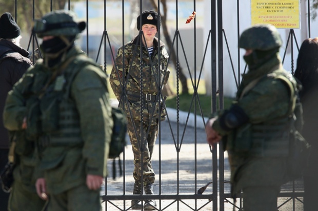 Một binh sĩ Ukraine đứng bên trong một căn cứ quân sự ở Crimea, trong khi các binh sĩ chưa xác định danh tính được trang bị vũ trang ở bên ngoài.