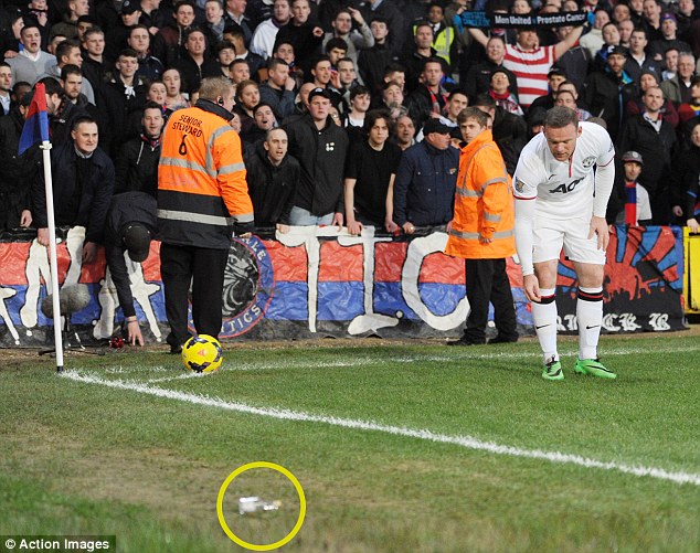Rooney cúi xuống nhặt đồng xu trong khi trên sân còn nhiều vật thể lạ khác bị ném xuống như một cái chai (khoanh tròn)