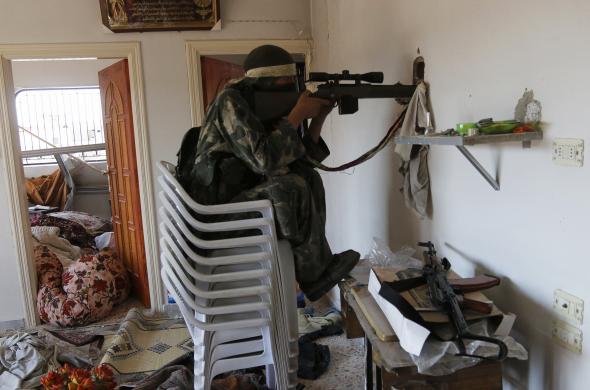 Một chiến binh phiến quân Syria ngồi trên ghế chĩa súng qua lỗ trên tường nhà tại thị trấn Morek, tỉnh Hama.