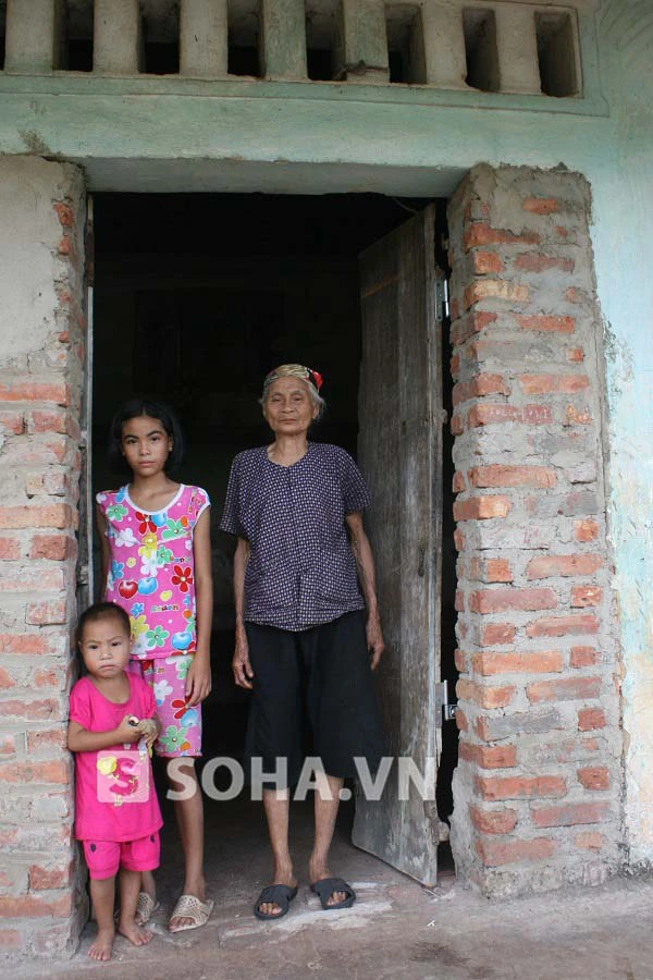 Bà Chỉ bên 2 đứa cháu nhỏ trước cửa ngôi nhà đã cũ nát của con trai bà