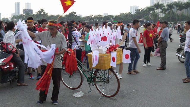 Một người đàn ông đang bán cờ cả Việt Nam và Nhật Bản. Cờ nhỏ giá 10 ngàn, giá tăng lên 20 ngàn và 30 ngàn với những cờ to hơn