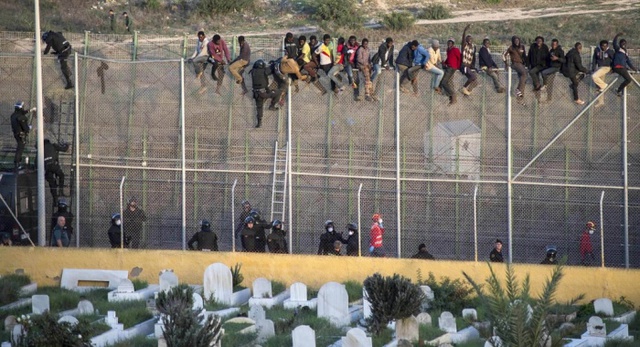 Người nhập cư châu Phi ngồi trên đỉnh hàng rào ngăn cách biên giới giữa Tây Ban Nha và Ma-rốc, trong khi cảnh sát Tây Ban Nha cố gắng đuổi người nhập cư trở tại phía Ma-rốc.