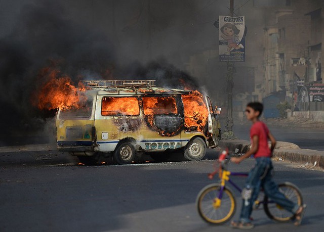 Một cậu bé dắt xe đạp qua chiếc ô tô đang bốc cháy trên đường phố ở Karachi, Pakistan.