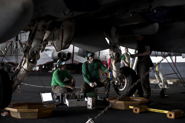 
Các thợ cơ khí, với màu áo xanh lá cây, đang bảo dưỡng bánh đáp của một chiếc máy bay trong nhà chứa trên tàu.
