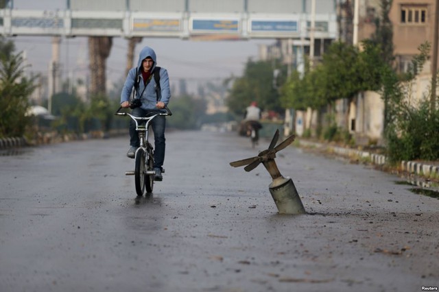 Người dân đạp xe qua một vỏ bom chùm găm xuống mặt đường ở thị trấn Douma, Syria.