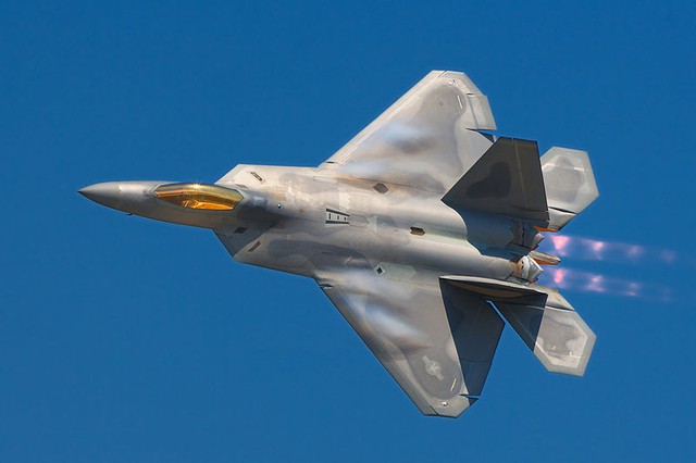 A pilot peers up from his F-22 Raptor while in-flight, showing the top view of the aircraft. The terrain of Nevada can be seen below mostly cloudless skies. Aircraft is mostly gray, apart from the dark cockpit windows.