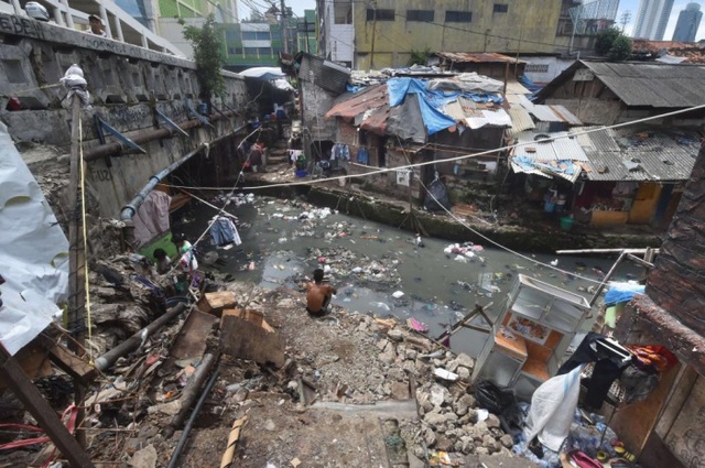 Người đàn ông cởi trần ngôi trên bờ sông gần những ngôi nhà ổ chuột tại thành phố Jakarta, Indonesia.