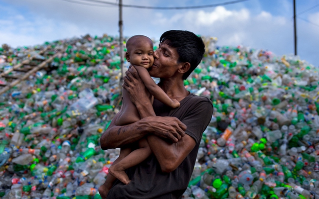 Người đàn ông bế con nhỏ đứng cạnh bãi thu gom đồ tái chế ở Dhaka, Bangladesh.