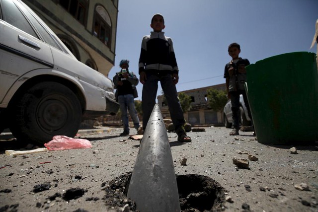 Các cậu bé đứng trước một quả đạn pháo chưa nổ găm xuống đường phố sau một đợt không kích vào thủ đô Sanaa, Yemen.