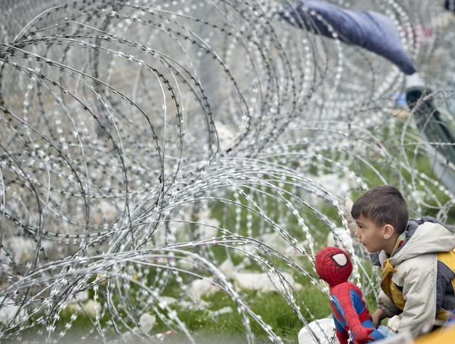 Cậu bé di cư chơi với búp bê cạnh hàng rào dây thép gai tại thị trấn Idomeni, Hi Lạp, giáp biên giới với Macedonia.