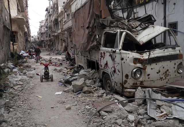 Bé gái đạp xe giữa những tòa nhà nổ nát và ô tô hỏng tại thành phố Homs, Syria.