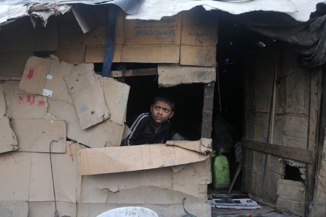 Cậu bé nhìn ra ngoài từ một túp lều tạm bằng bìa cát tông ở Srinagar, Ấn Độ.