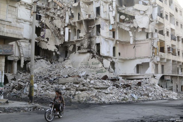 Mọi người đi xe máy qua những tòa nhà đổ nát do nội chiến ở Aleppo, Syria.