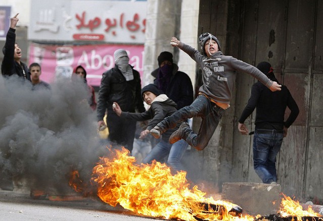 Một cậu bé người Palestin ném đá vào binh sĩ Israel khi nhảy qua lốp xe đang bốc cháy trong cuộc đụng độ ở Hebron, Bờ Tây.