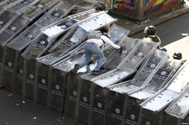 Người biểu tình trèo lên hàng rào khiên của cảnh sát chống bạo động ở thủ đô Caracas, Venezuela.